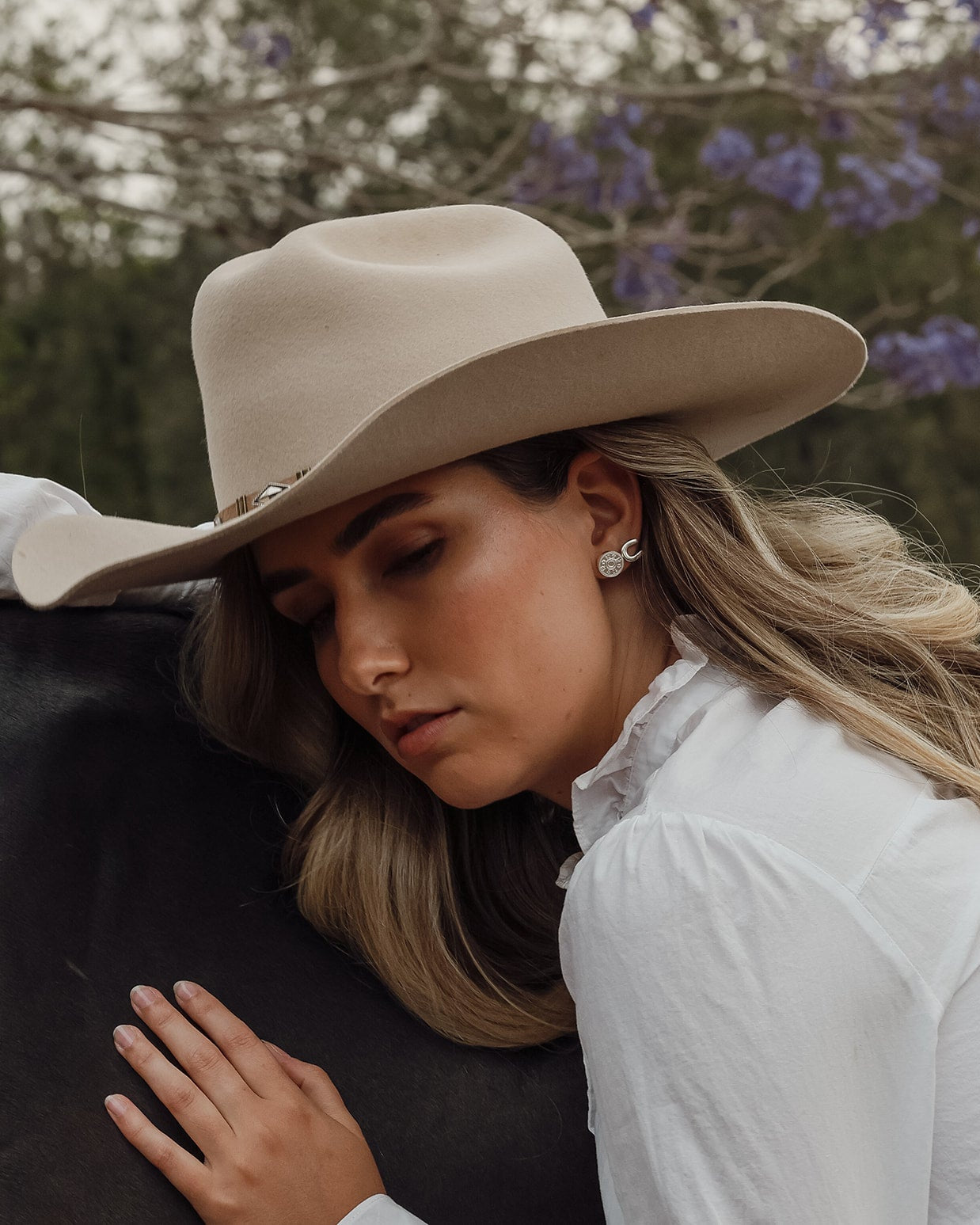 A woman wearing a wide-brimmed beige cowboy hat and a white blouse rests her head on a dark blanket outdoors, with blurred trees and purple flowers in the background.