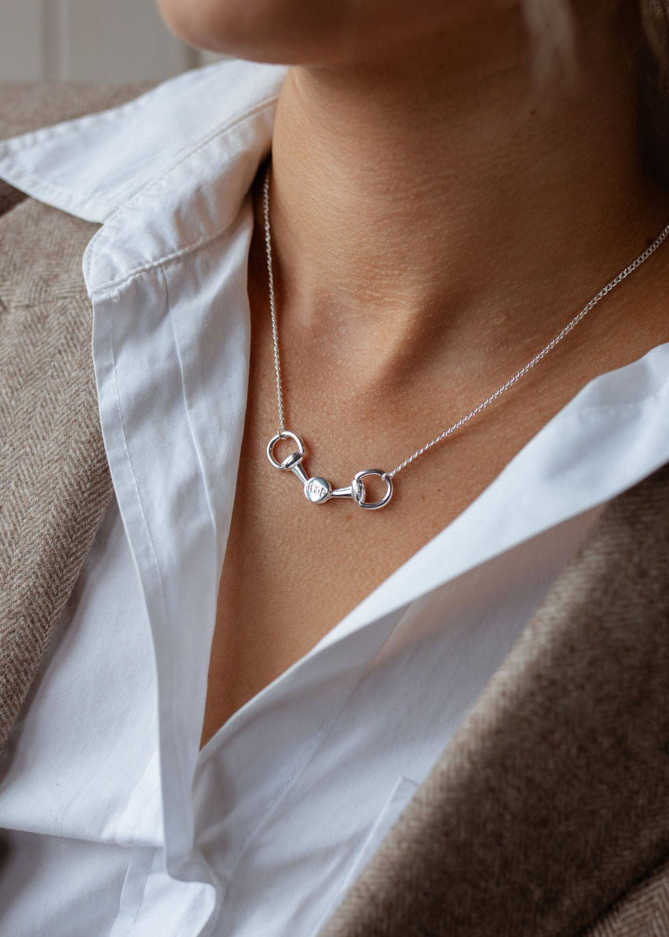 A close-up of a woman wearing a delicate silver necklace with a horse bit pendant, styled over a white collared shirt and a brown blazer.