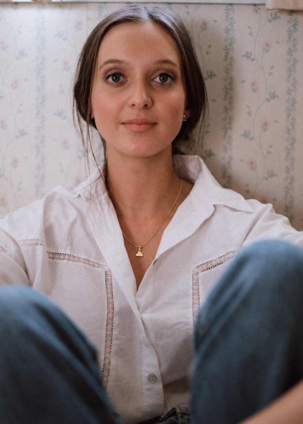 A woman wearing a white button-up shirt with delicate gold embroidery on the chest and a small gold pendant necklace, sitting against a vintage floral wallpaper background.
