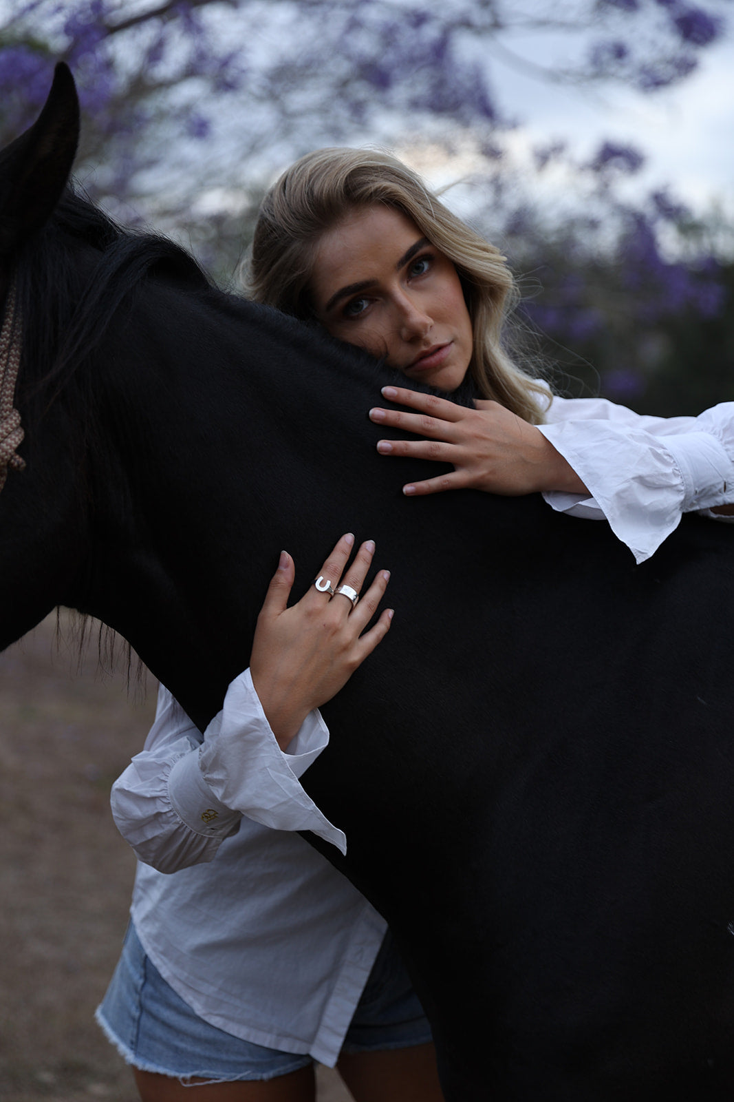 A woman in a white blouse and denim shorts poses closely with a black horse outdoors, wearing a silver ring on one finger. Purple flowering trees are blurred in the background.
