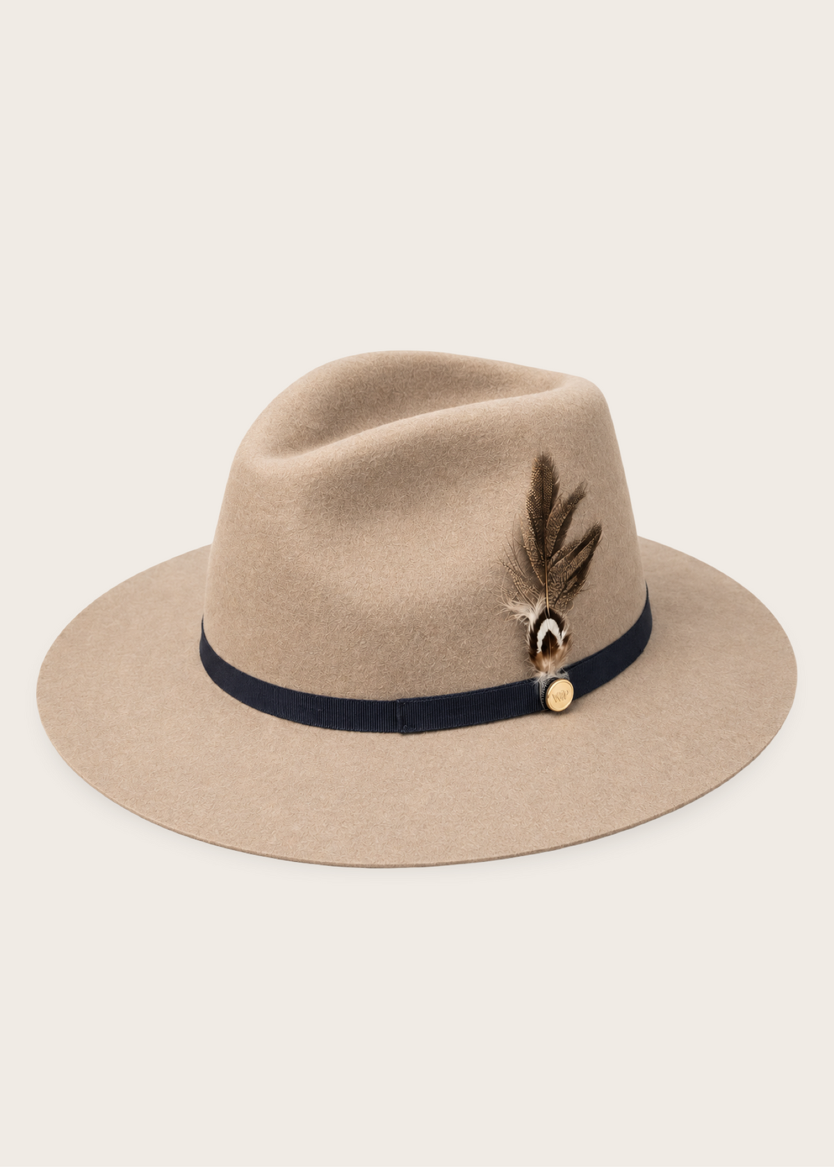 A beige felt fedora hat with a dark navy band, a brown feather, and a small gold button on the side, photographed against a solid off-white background.