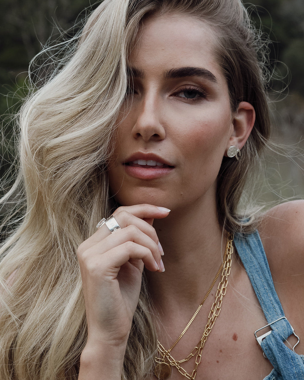 A close-up lifestyle shot of a woman with long blonde hair wearing layered gold necklaces, a gold ring, and a silver stud earring, paired with a denim top.