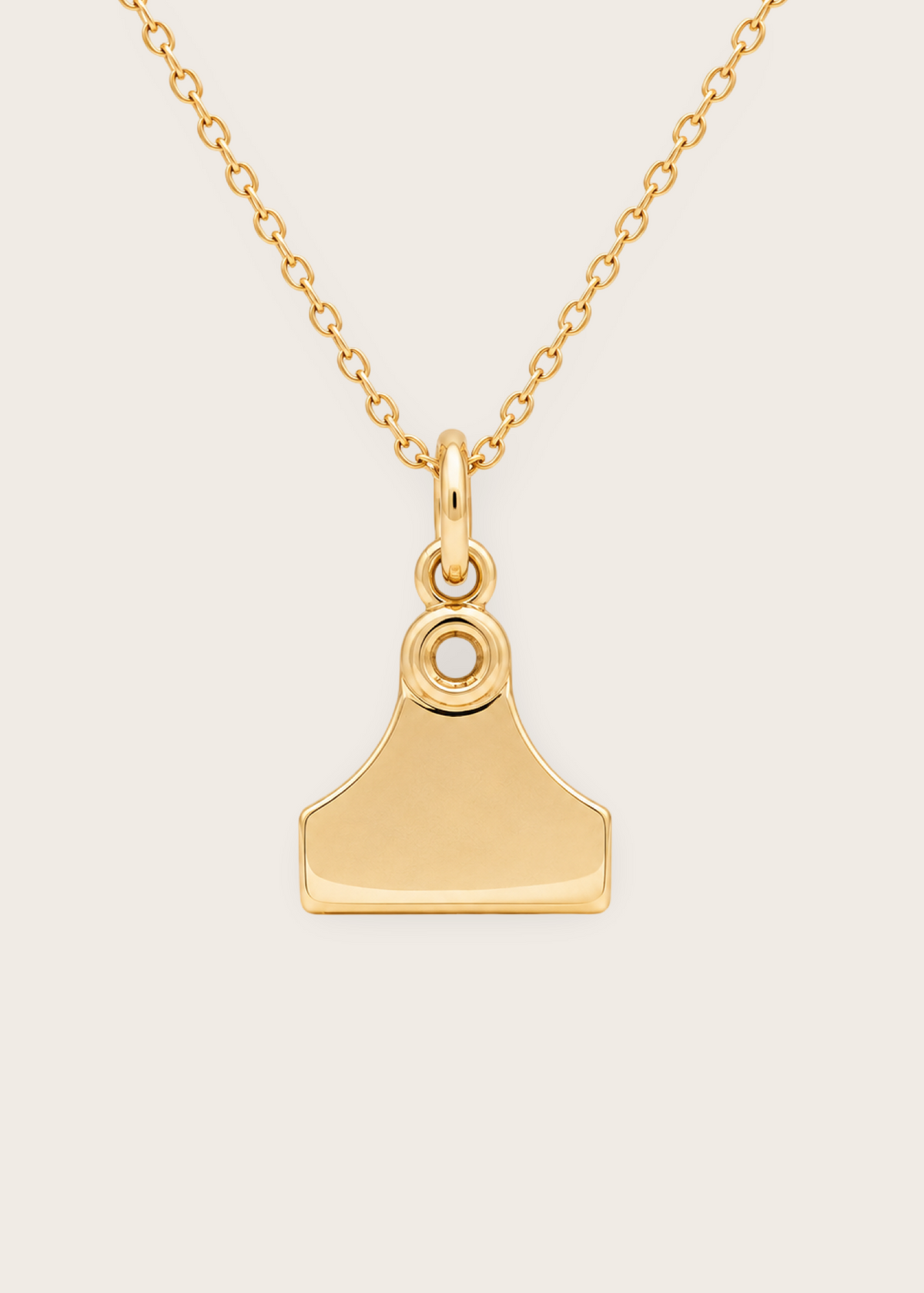 A gold pendant necklace featuring a minimalist, shield-like or house-shaped charm with a small circular detail at the top, suspended from a delicate gold chain against a light beige background.