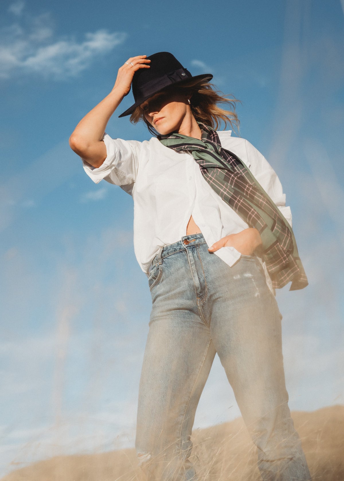 A woman wearing a navy wide-brim hat, white button-up shirt, and light blue jeans stands outdoors in a field under a blue sky, holding her hat with one hand and wearing a plaid scarf.